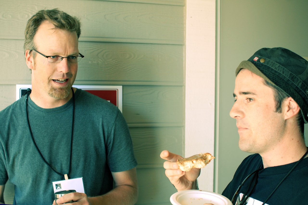 Two men standing and talking; one holds a drink, the other eats while listening.