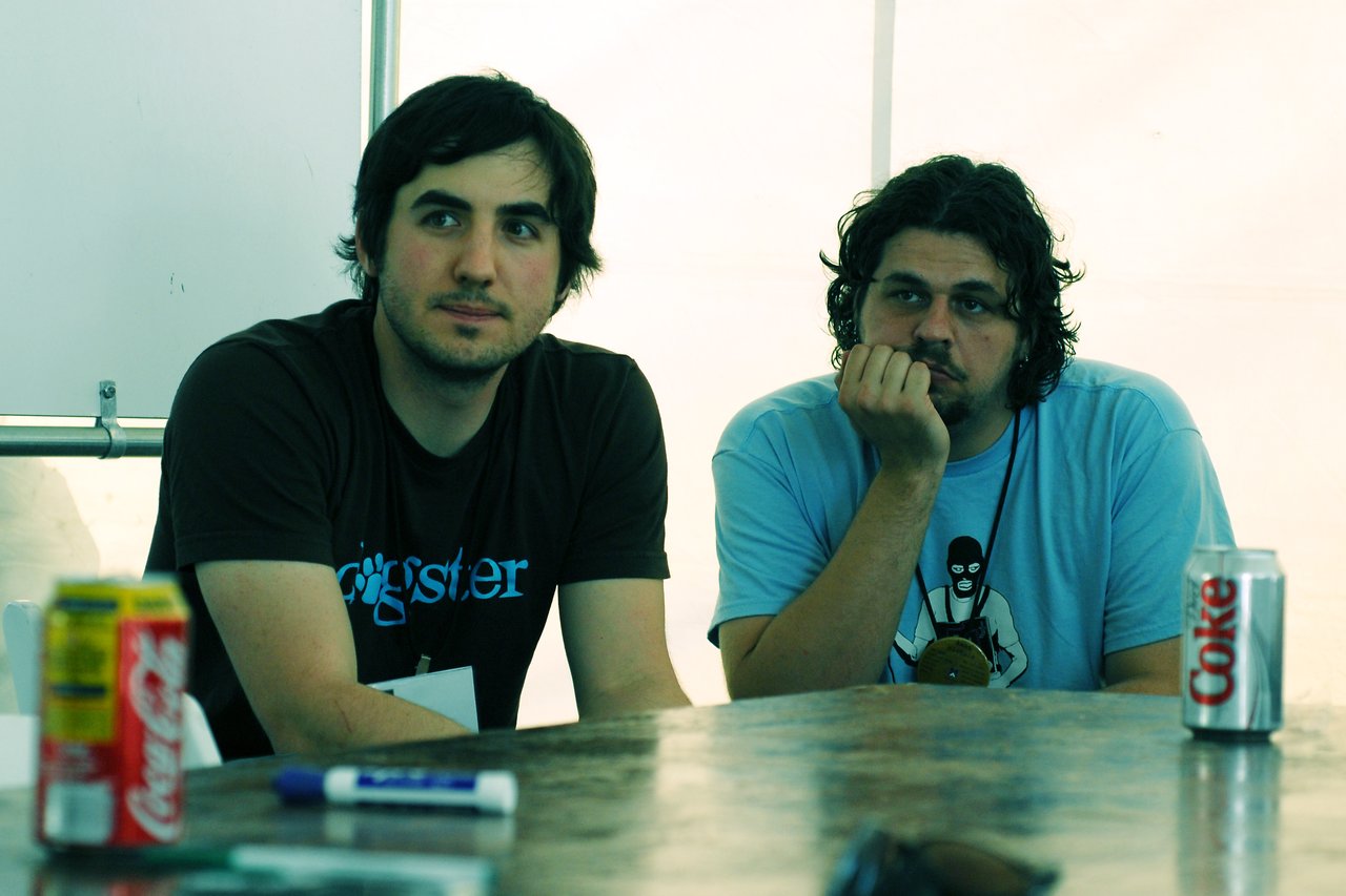 Two men sit at a table, one attentively listening while the other rests his chin on his hand.