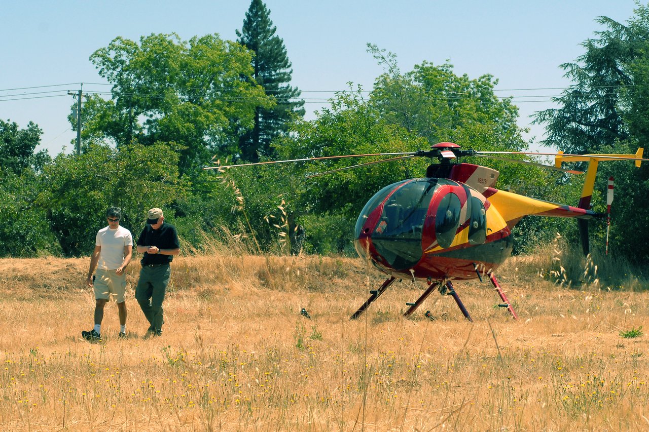 Two people walk away from a colorful helicopter parked in a dry grassy field.