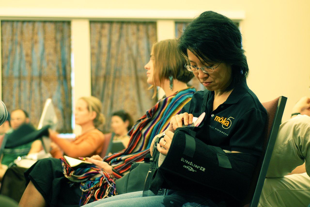 A woman wearing glasses and a black polo shirt sits in a conference room, adjusting her arm sling.