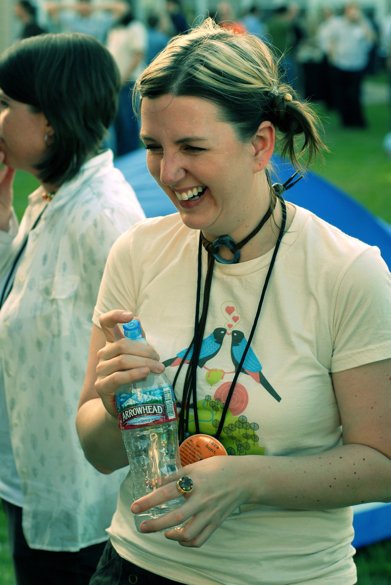 A smiling person with short, tied-back hair holds a water bottle at an outdoor event, wearing a graphic t-shirt.