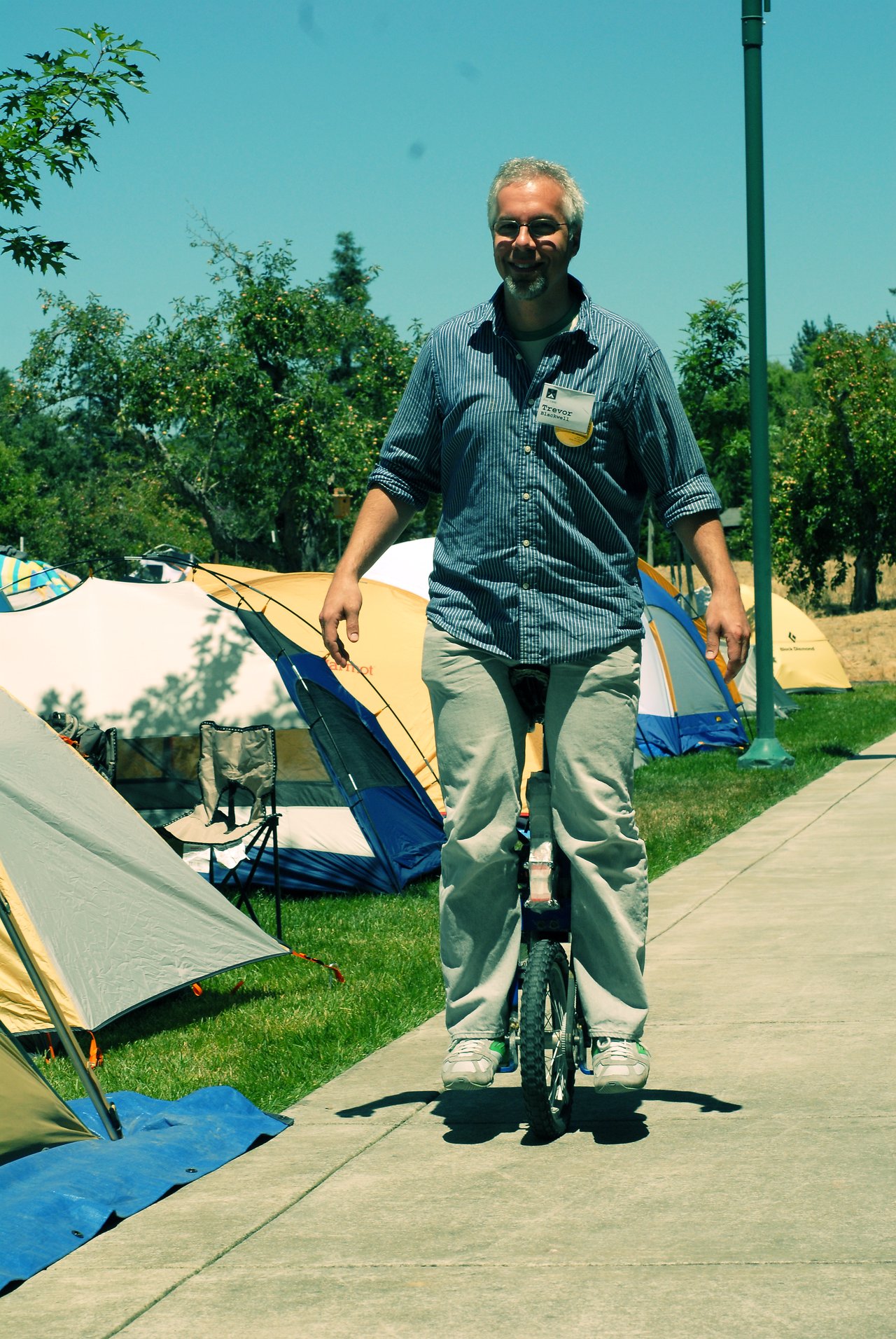 A man rides a unicycle on a paved path, smiling, with tents and camping gear in the background.