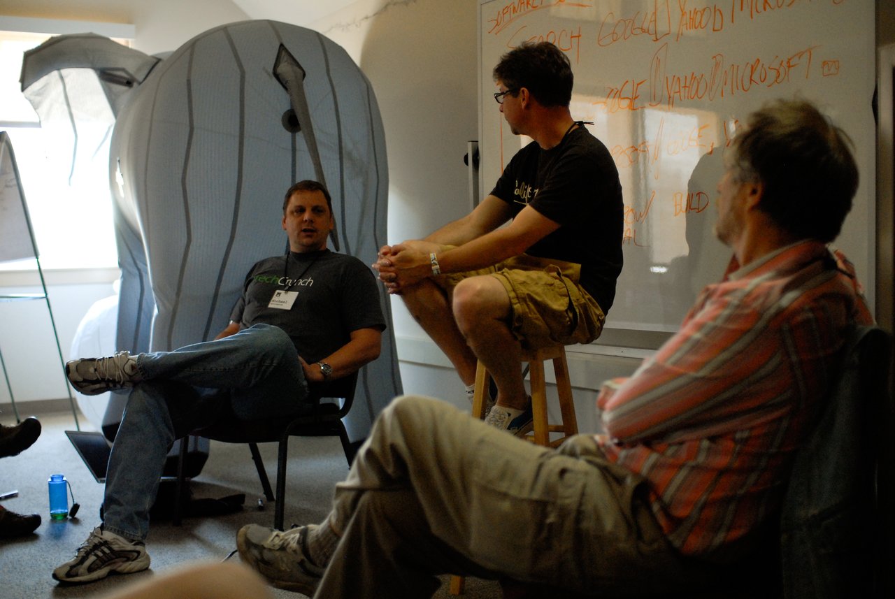 Two men engage in a debate about search engines while a moderator and others listen in a discussion setting.