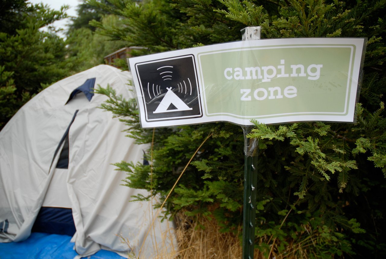 A "camping zone" sign is attached to a post near a tent, surrounded by green bushes.