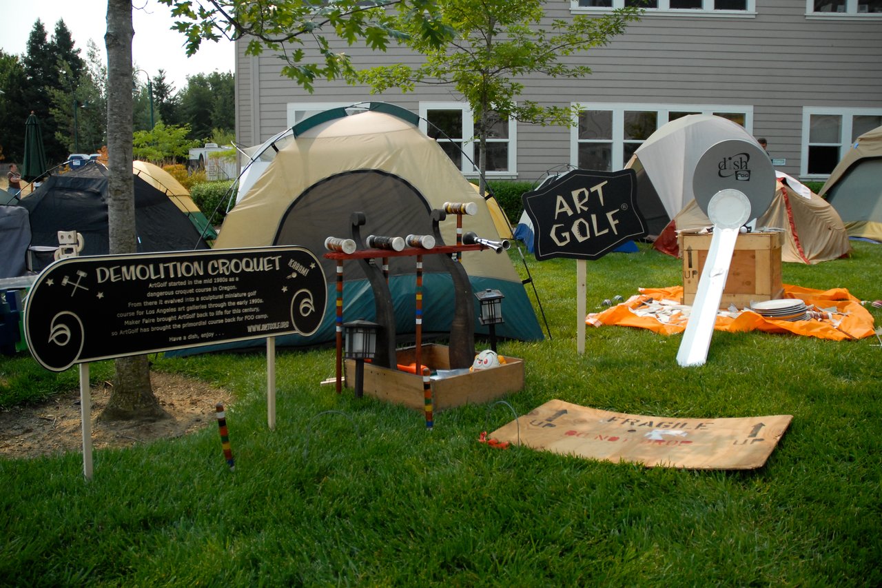 Outdoor setup with tents and signs for "Demolition Croquet" and "Art Golf", featuring creative game equipment on grass.