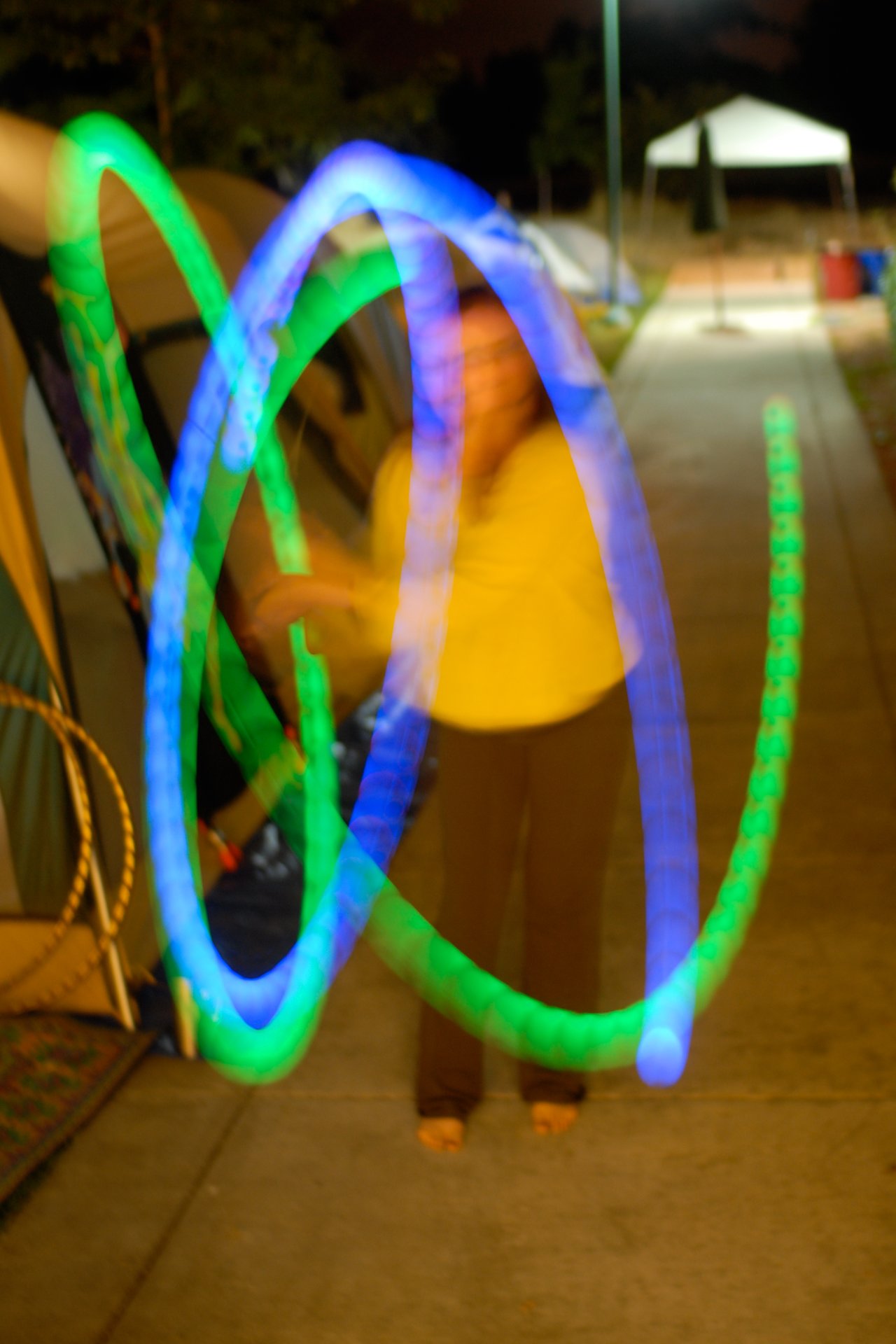 A person in a yellow shirt moves glowing blue and green light trails in a blurred motion at night.