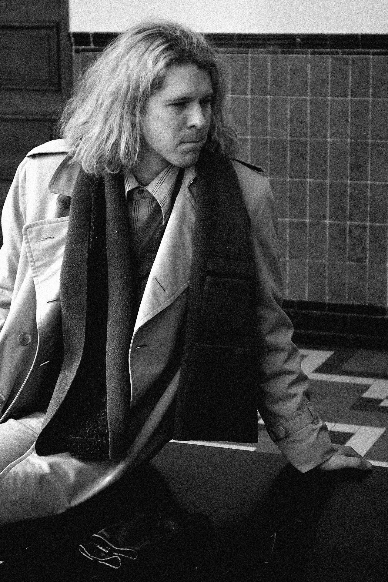 A person with long hair, wearing a trench coat and scarf, sits on a table and looks to the side.
