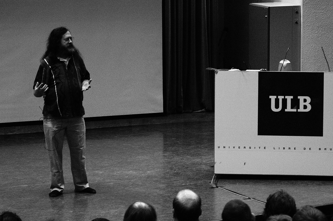 A speaker with long hair and a beard gestures while giving a talk on stage at a conference.