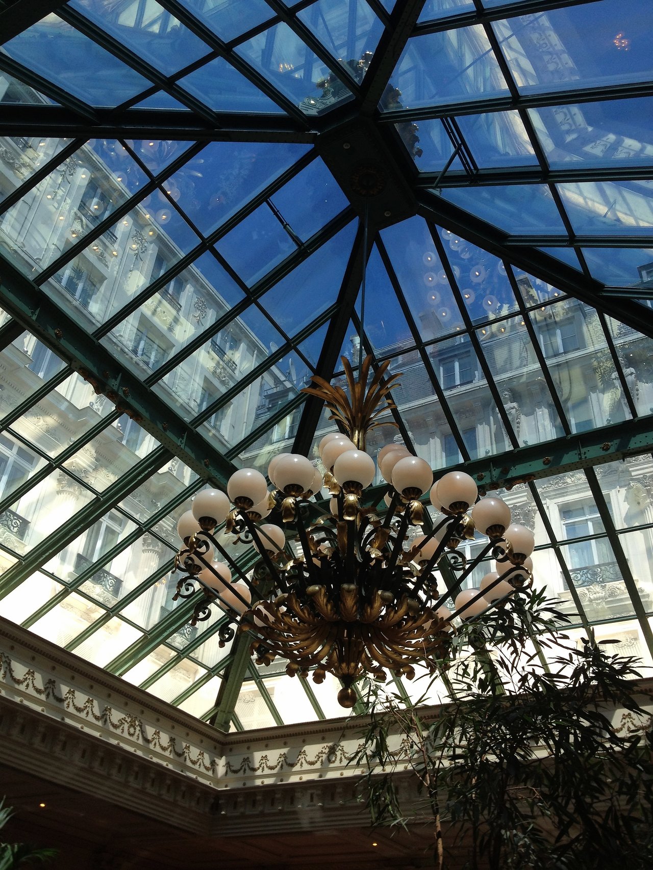 A large ornate chandelier hangs under a glass ceiling, reflecting surrounding buildings at Café de l'Opéra in Paris.