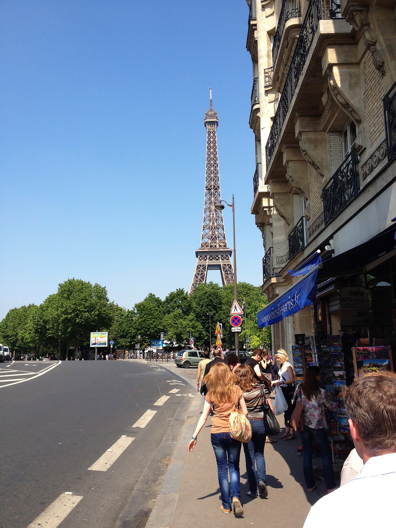 People walking on a sidewalk with the Eiffel Tower in the background on a sunny day in Paris.