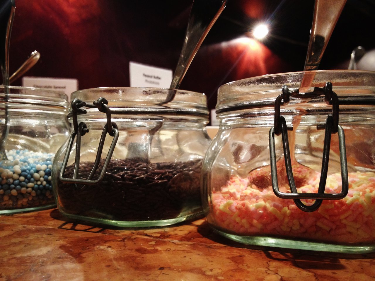 Three glass jars filled with Dutch sprinkles, including chocolate hagelslag and colorful muisjes, with spoons inside.