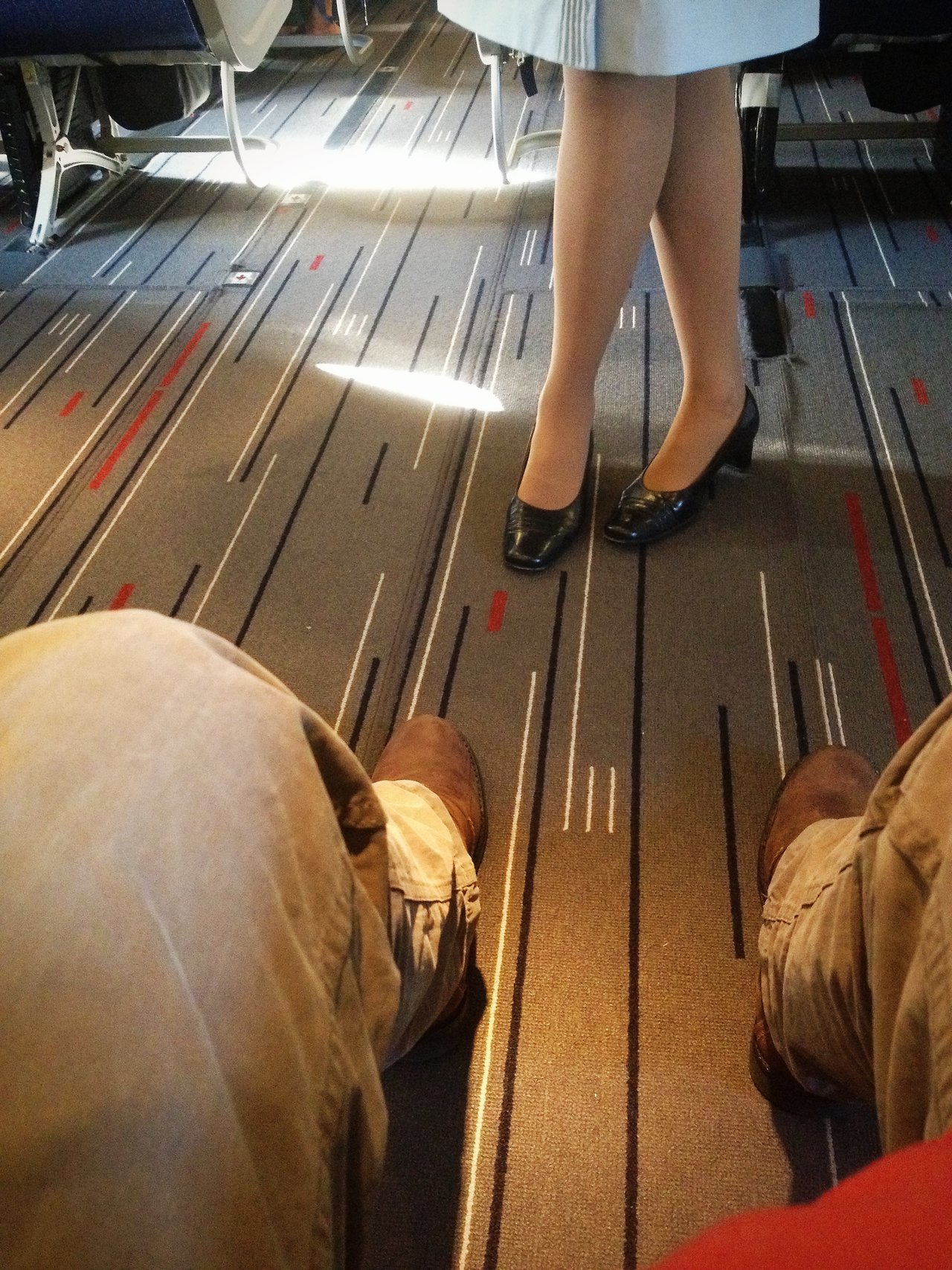 A seated passenger's legs stretch out in front of them, facing a standing flight attendant in a light blue uniform.