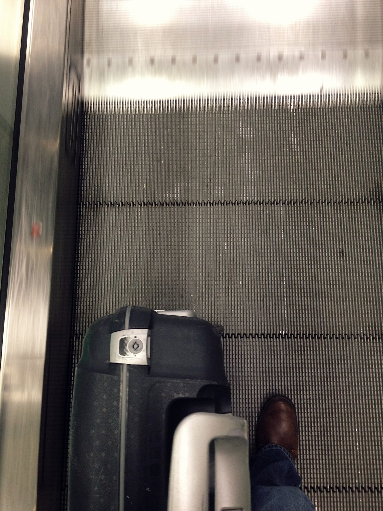 A person with a suitcase stands on a moving walkway, wearing jeans and brown shoes.