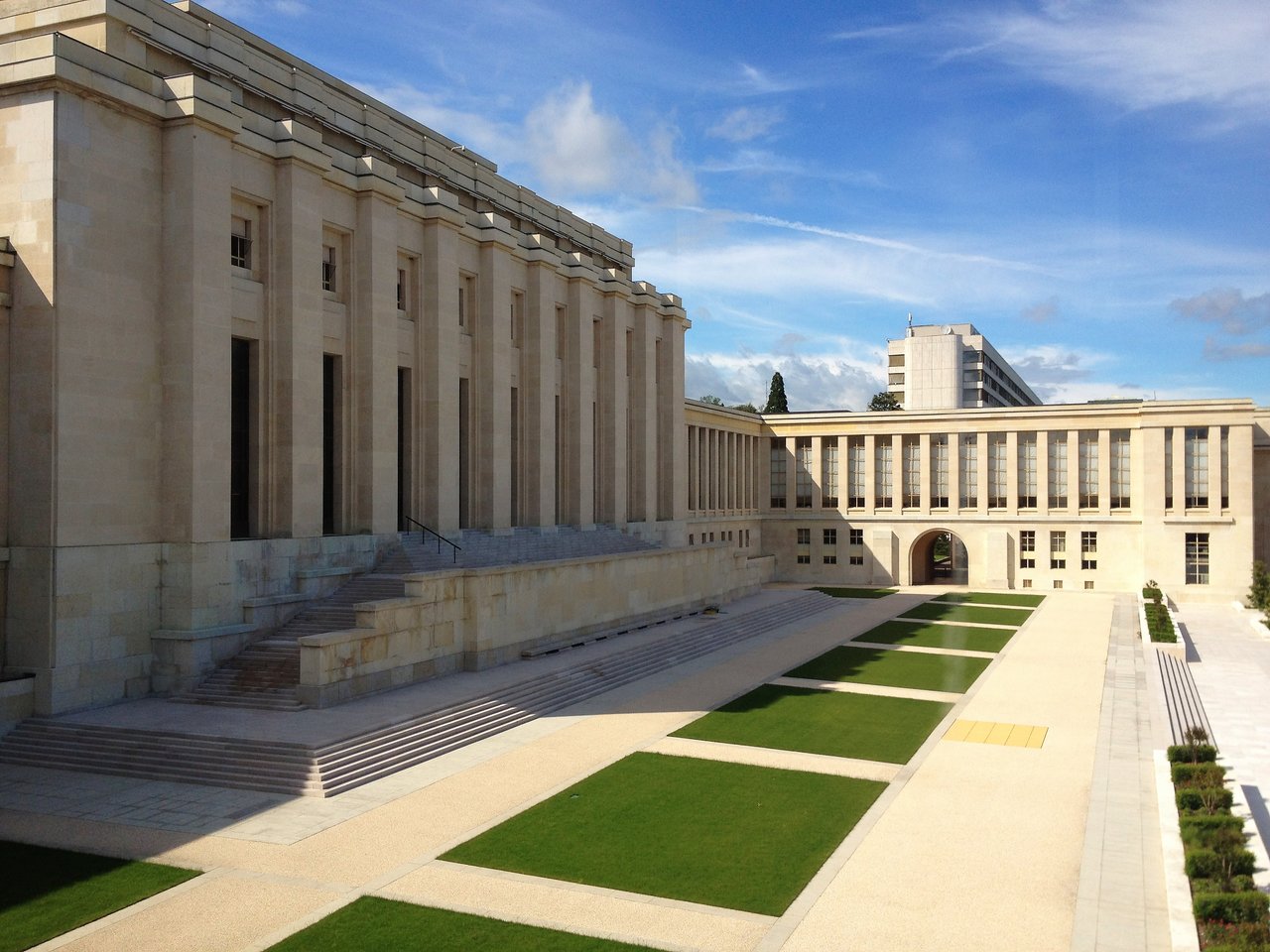 The Palais des Nations in Geneva, a large beige stone building with columns, green lawns, and a clear blue sky.