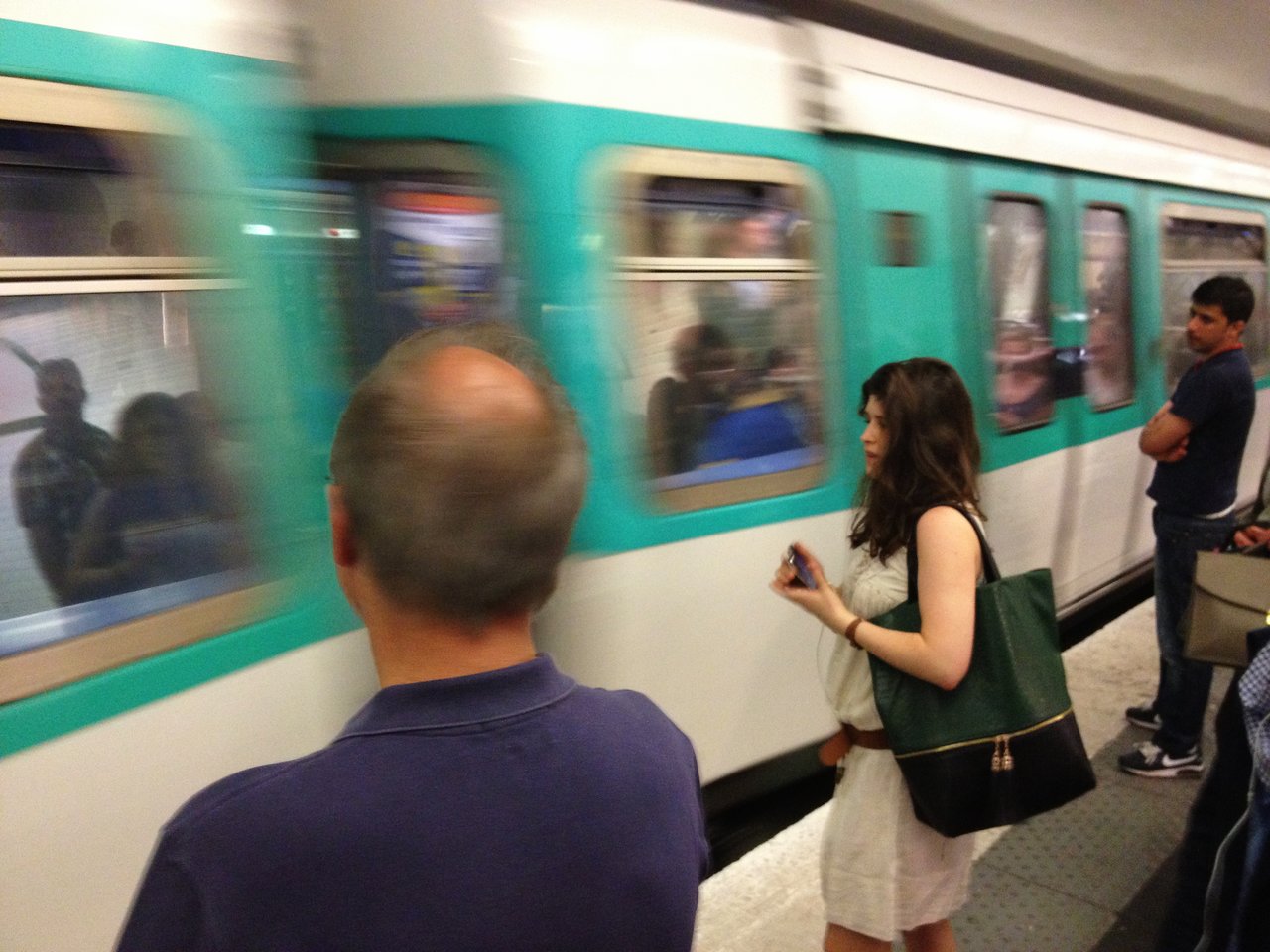 A metro train arrives at the platform as passengers wait, including a woman holding a phone and a large bag.