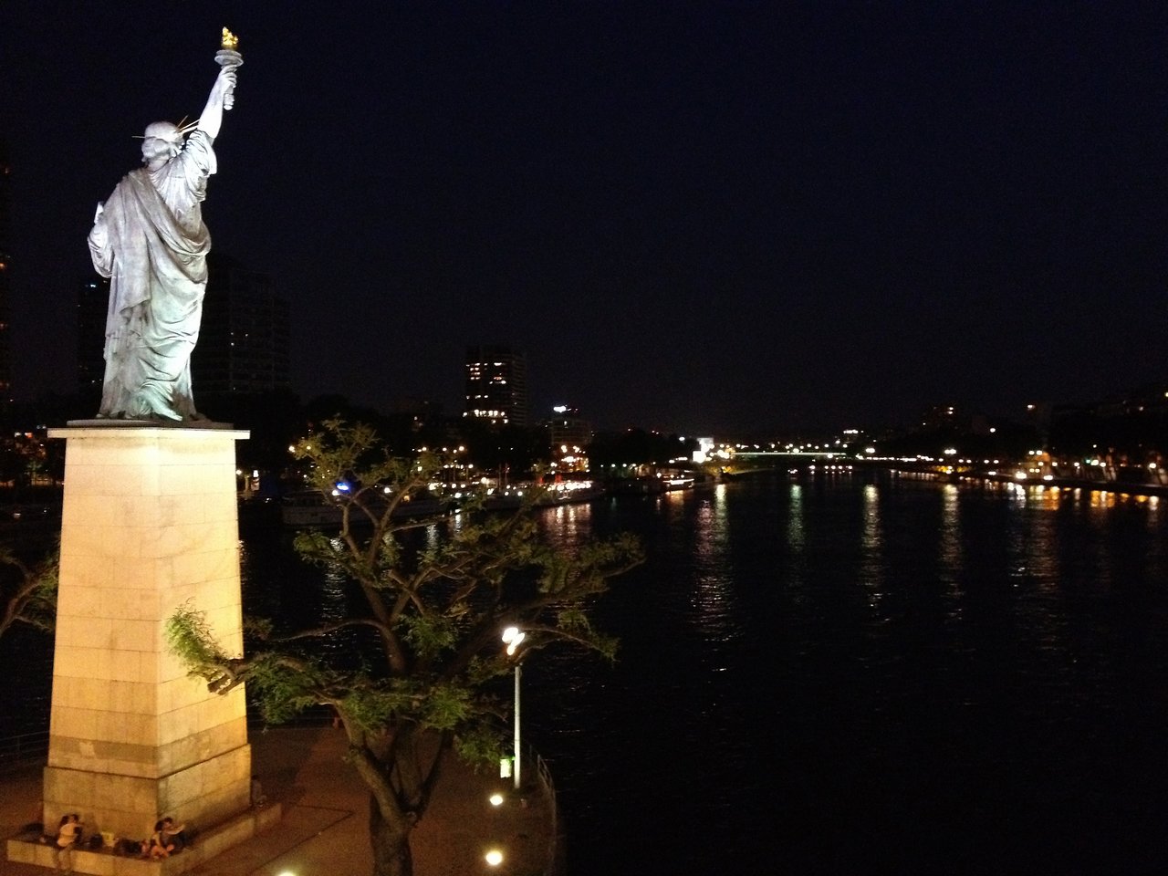 Statue of Liberty replica in Paris at night, standing on a pedestal near the Seine River with city lights.
