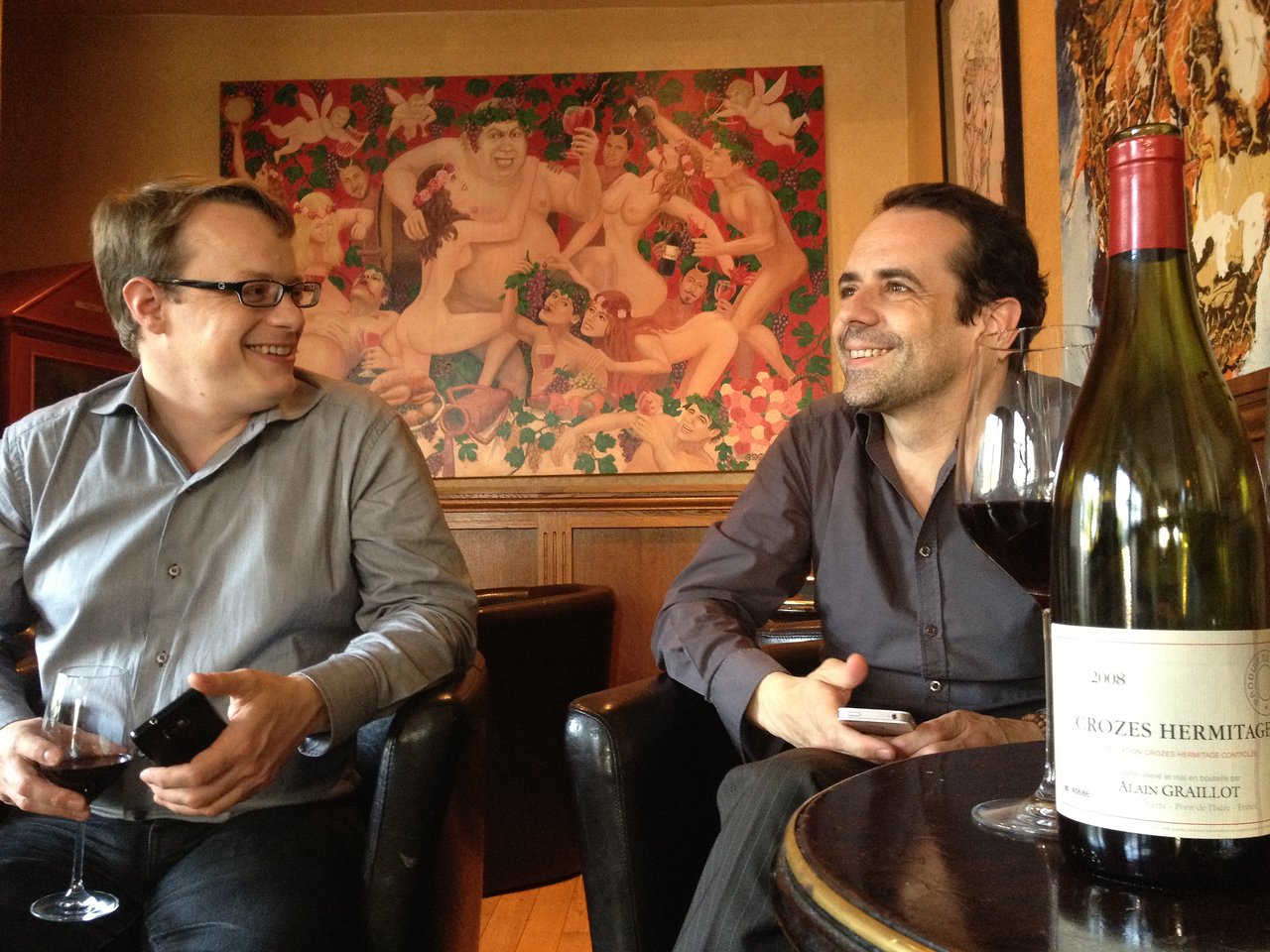 Two men sitting in a bar, smiling and holding wine glasses, with a bottle of red wine on the table.