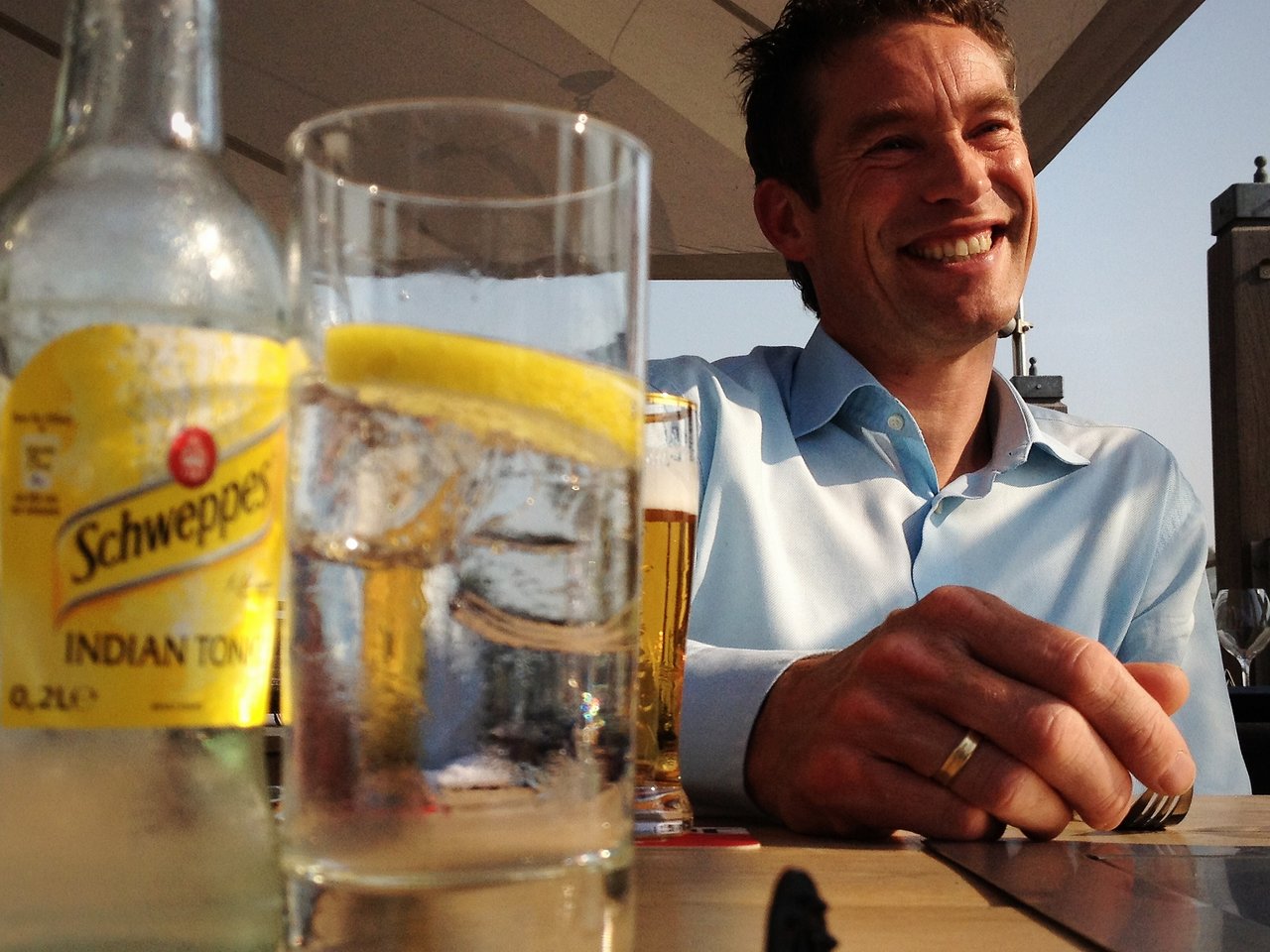 A smiling man in a blue shirt sits at an outdoor table with drinks, including tonic water and beer.
