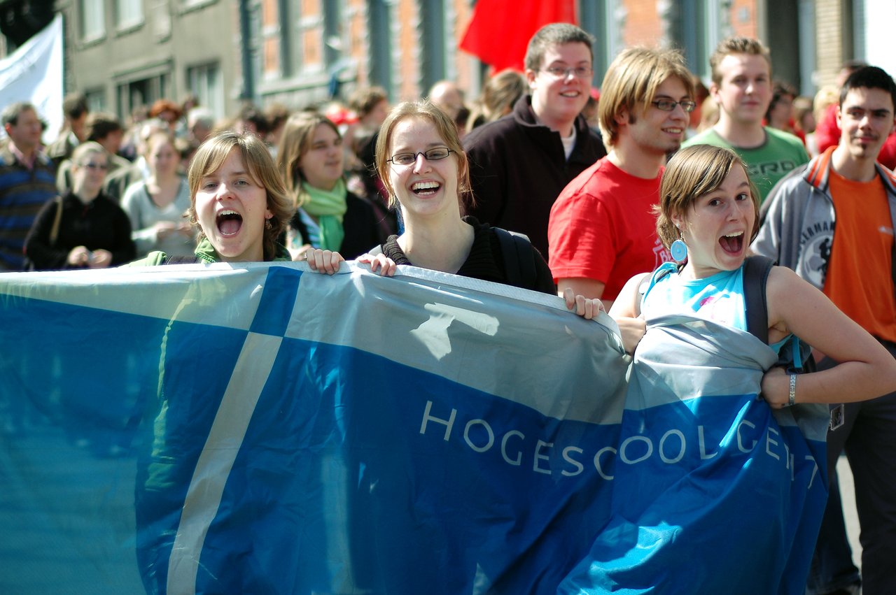 A group of young people hold a "Hogeschool Gent" banner, smiling and shouting during a protest or demonstration.
