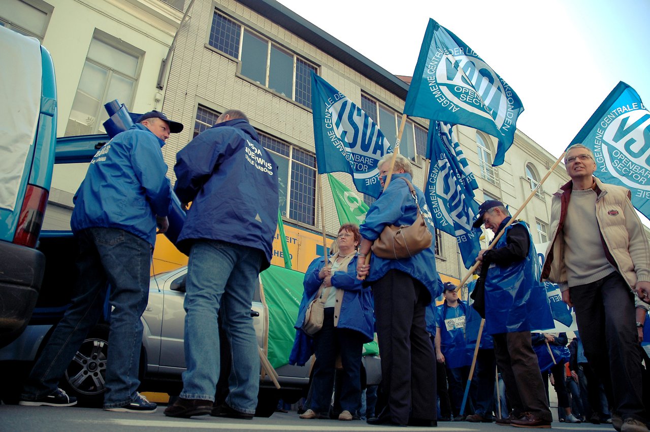 Protesters in blue jackets hold flags and gather on a city street during a demonstration.