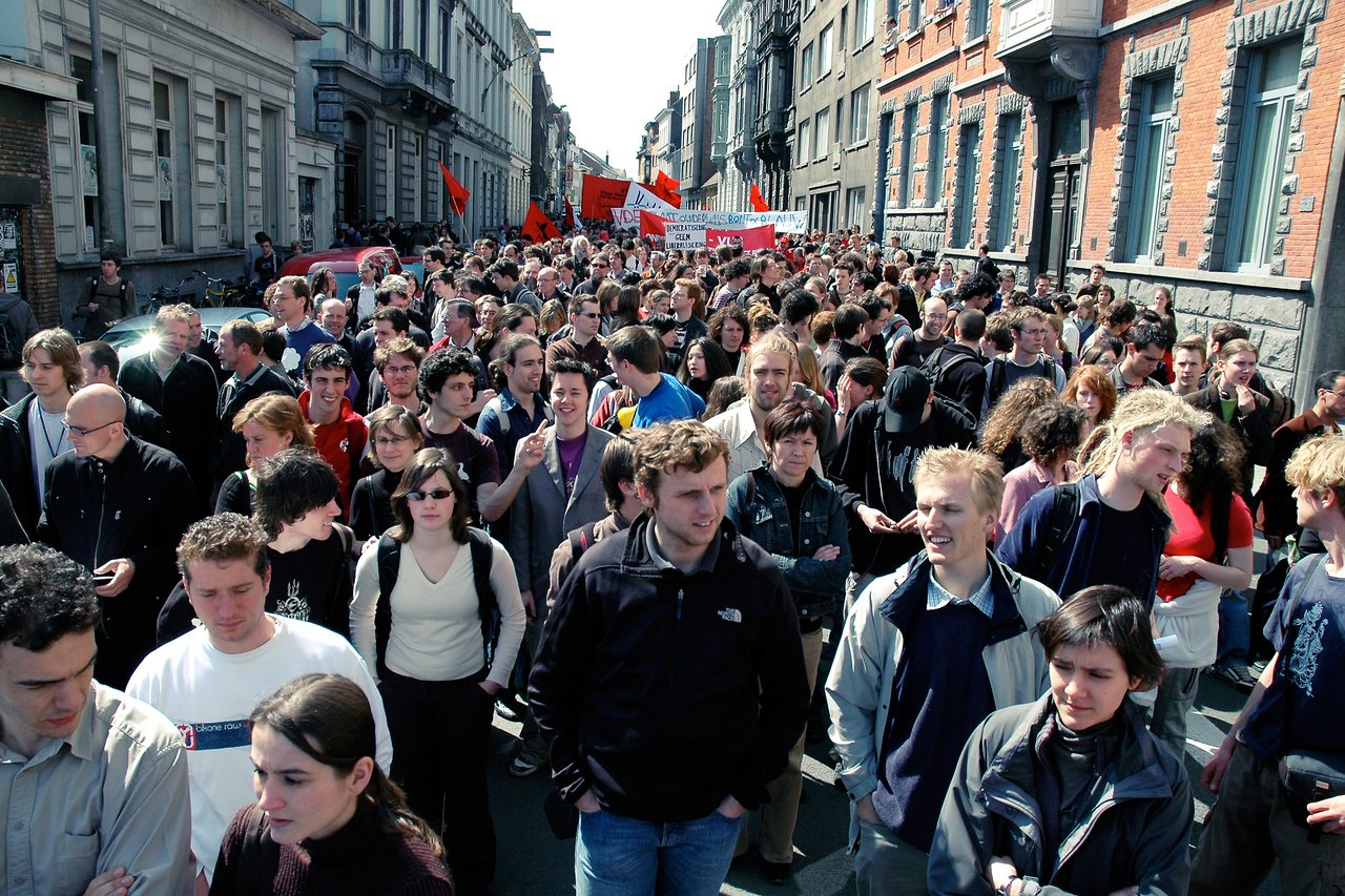 A large crowd marches down a city street, holding red flags and protest signs.