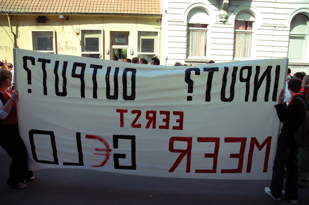 Protesters stand in the street holding a large banner with bold, reversed text during a demonstration.