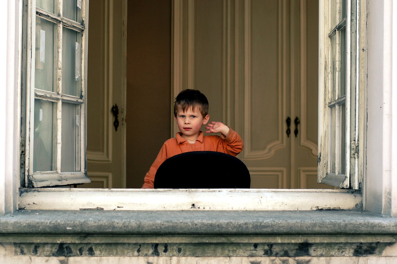 A young child in an orange shirt looks out an open window, holding one hand to their ear.
