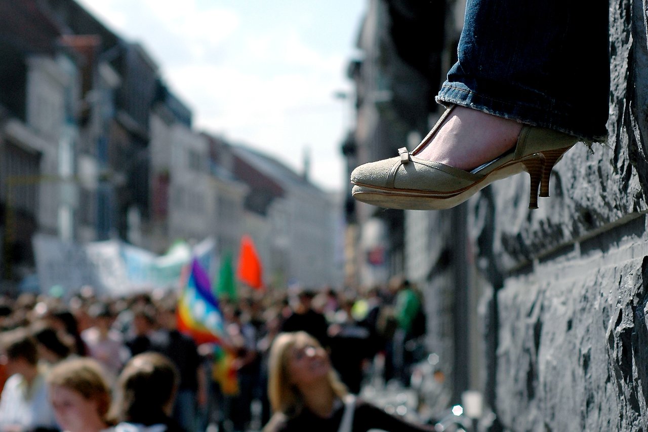 A person sits on a ledge with their feet dangling, watching a crowded protest with colorful flags below.