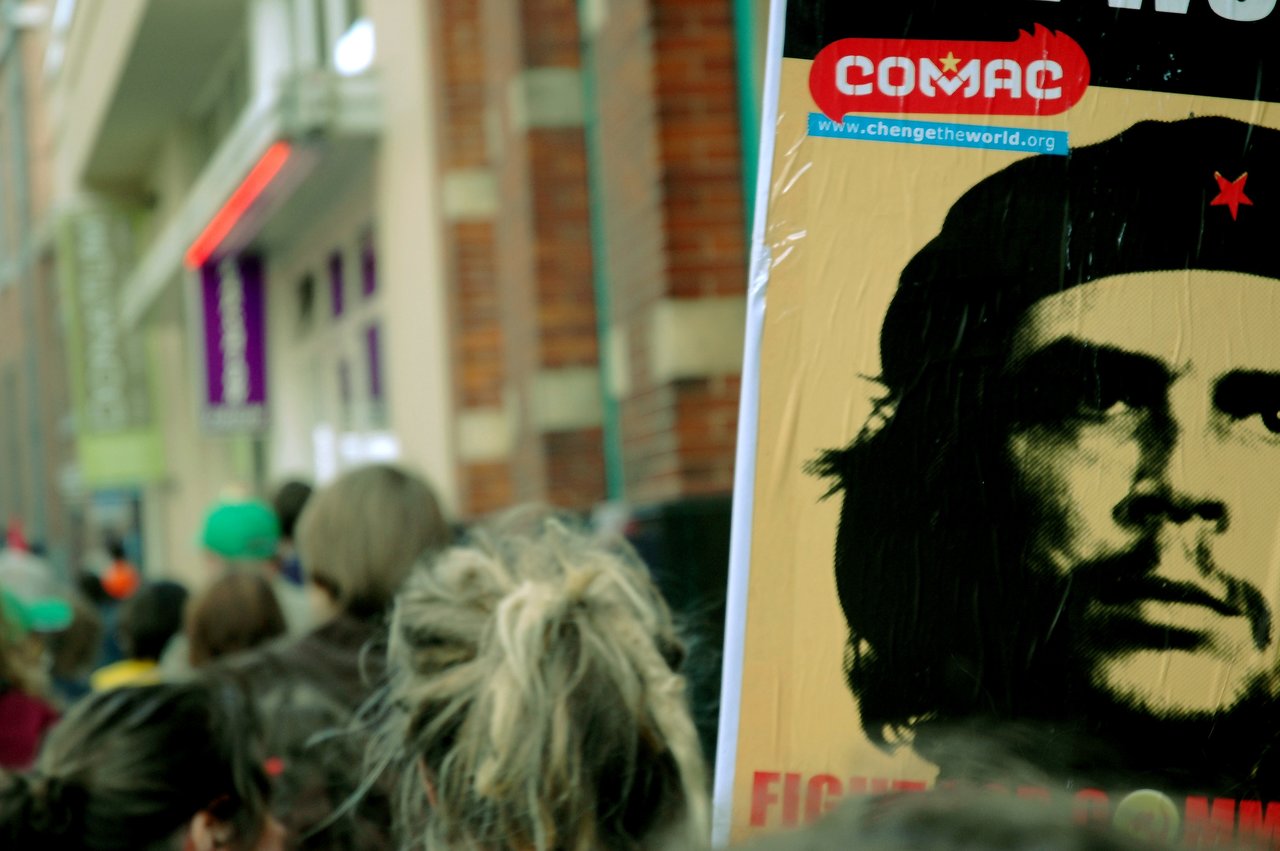 A protest crowd marches, holding a large sign with Che Guevara's portrait and the word "COMAC.