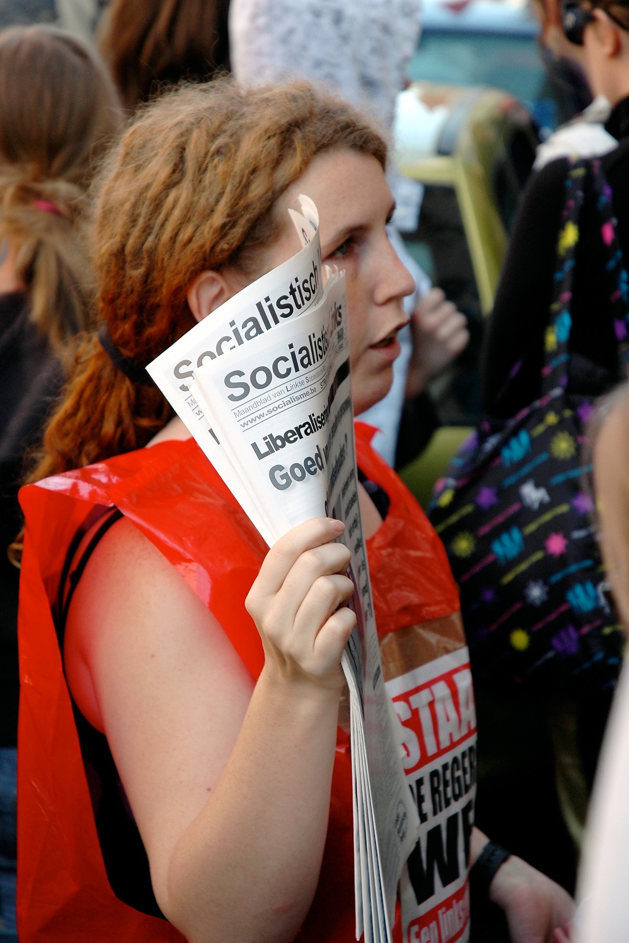A protester in a red vest holds and distributes socialist newspapers during a demonstration in a crowded public space.