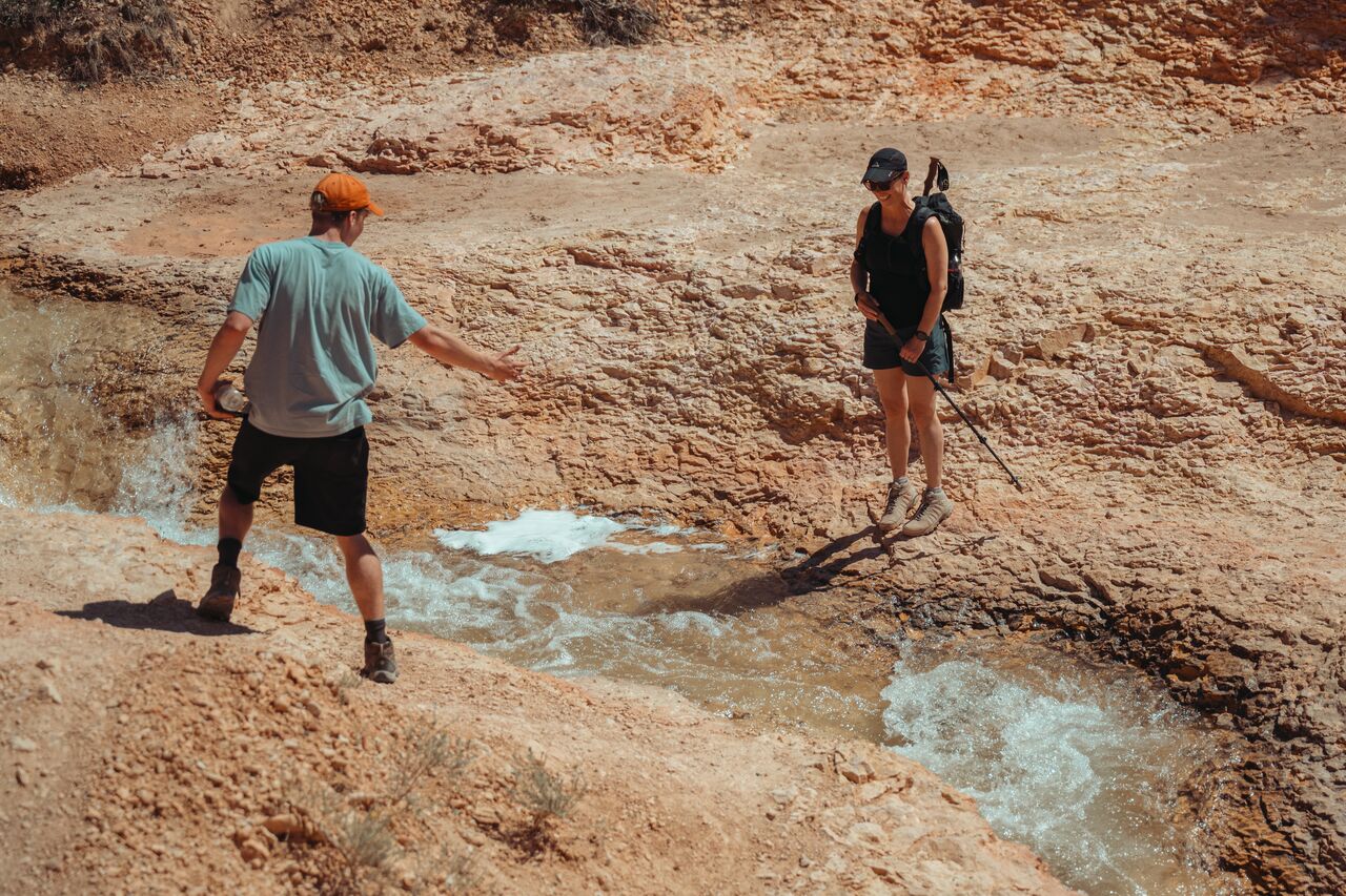 A young man offering a hand to a woman preparing to jump across a narrow stream on a rocky trail in Bryce Canyon.