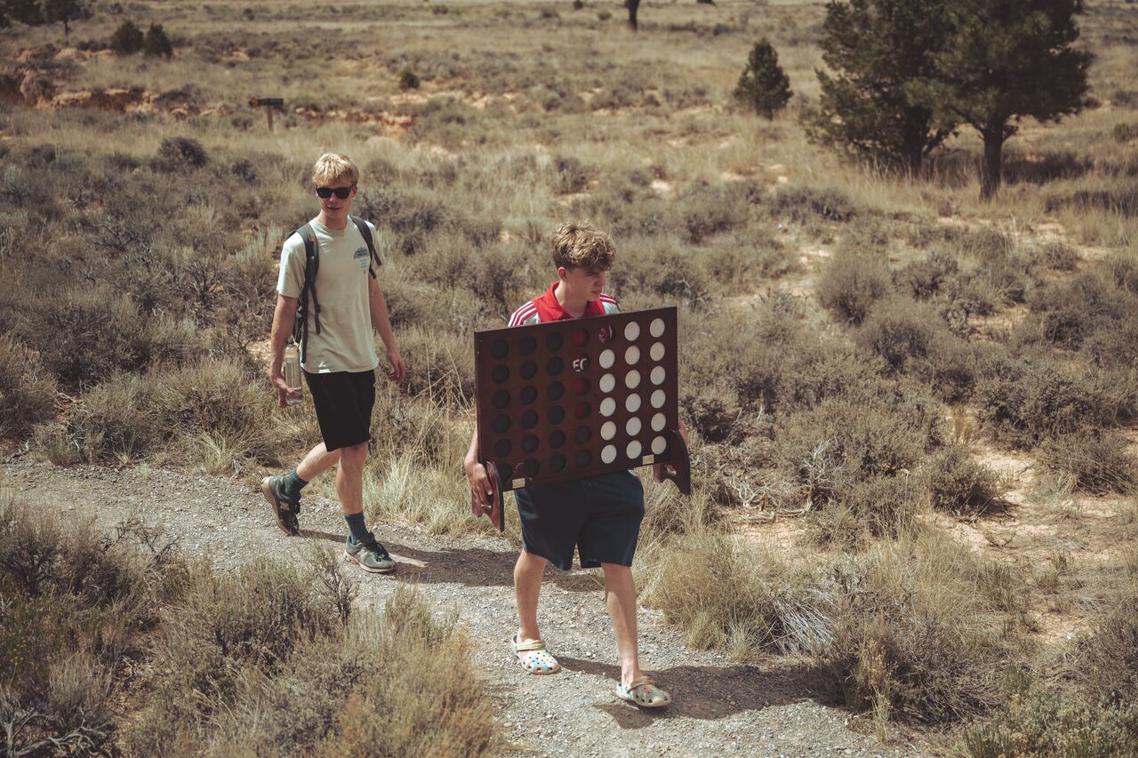 A young adult carrying a large Connect Four board on a desert trail with another young adult behind him.