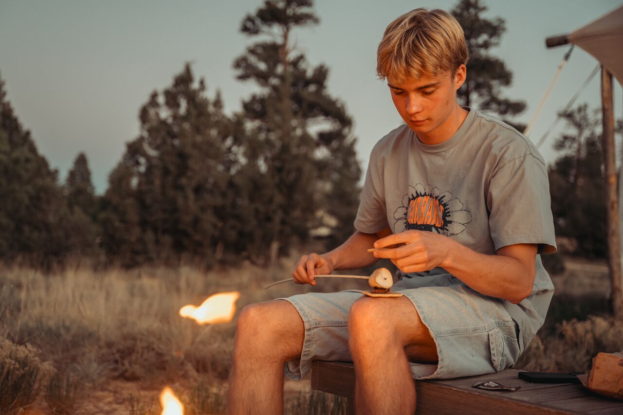 A young adult sitting by a fire pit, taking a roasted marshmallow off a stick and placing it onto a cookie on his knee.