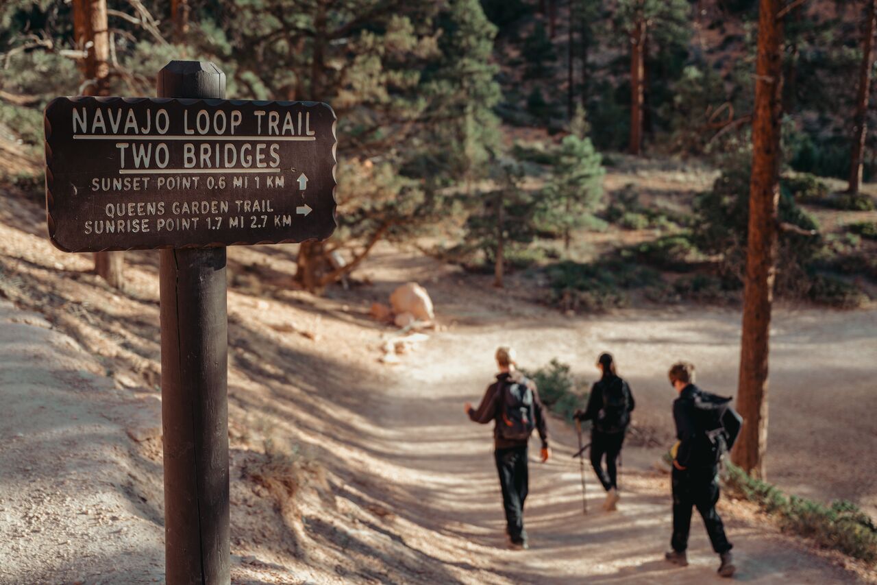 Three hikers starting down the Navajo Loop Trail past a trail sign in Bryce Canyon National Park.