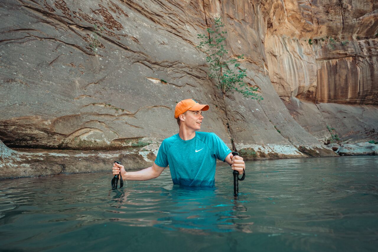 A young person wades through waist-deep water using trekking poles for balance.