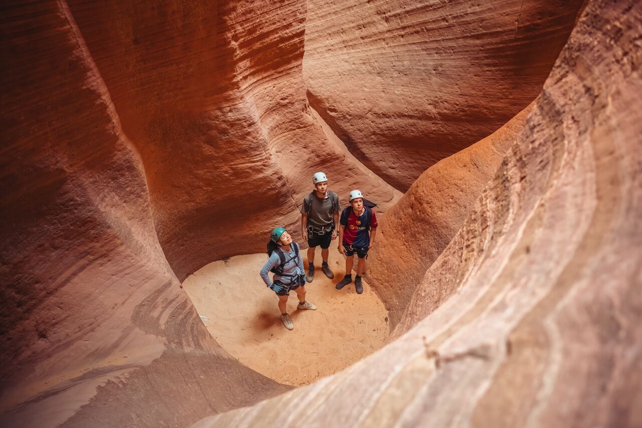 Three people wearing helmets standing at the bottom of a sandstone chamber in Zion National Park, looking up at the canyon walls.