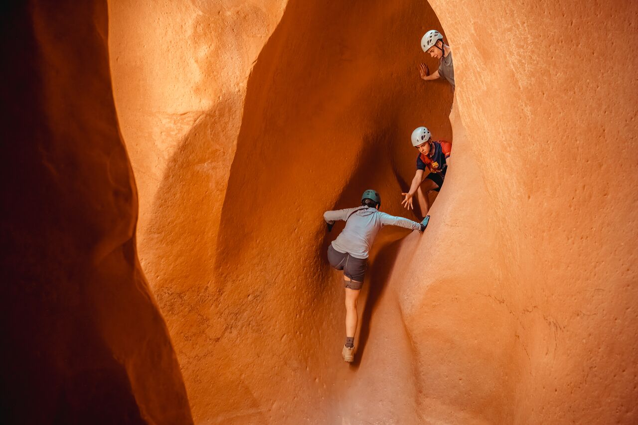 Two young adults reaching down to help a parent climb up a steep sandstone wall in Zion National Park.