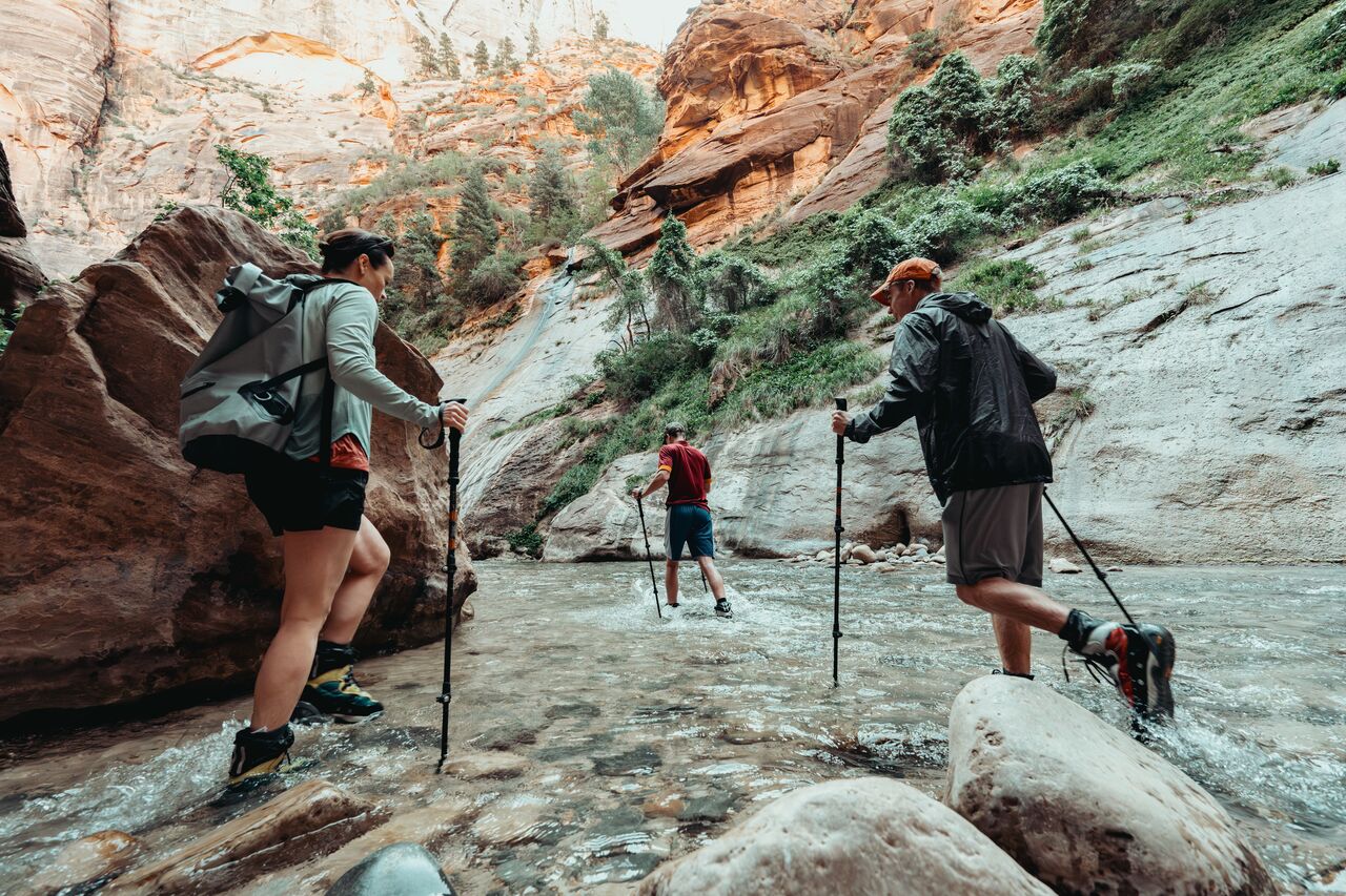 Three people walking up the river with their boots in the water in the Narrows at Zion National Park.