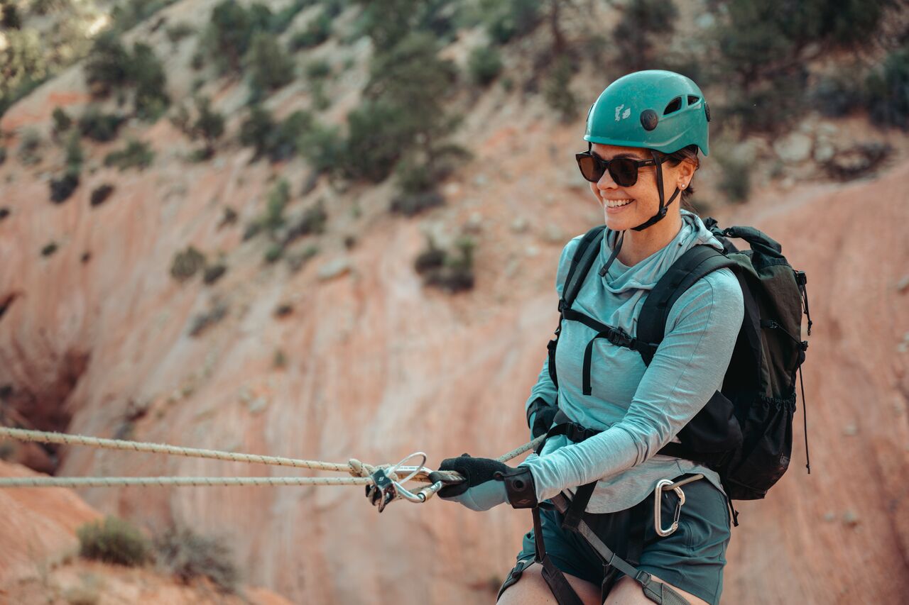 A woman wearing a helmet and sunglasses, smiling while rappelling in Zion National Park.