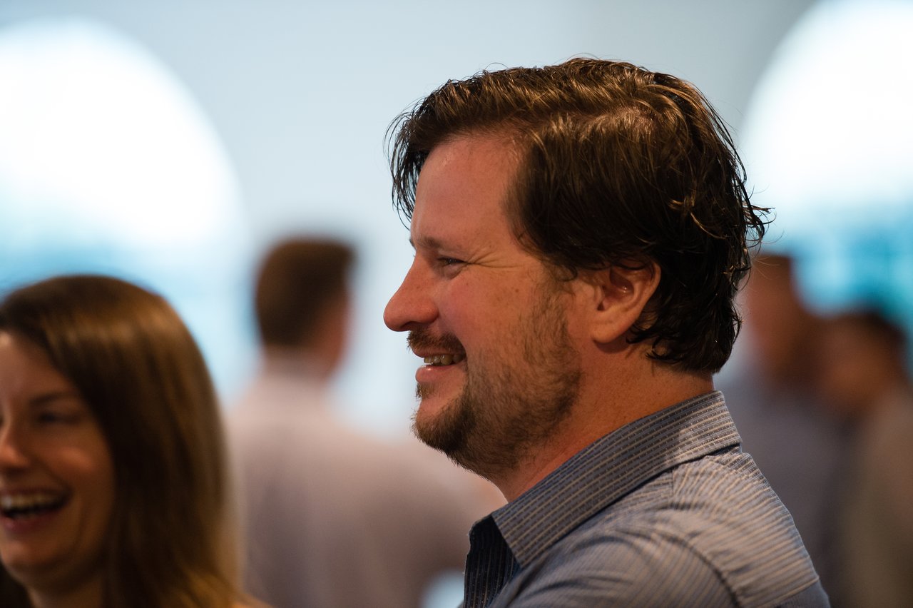 A man in a blue shirt smiles while attending an awards dinner with other guests in the background.