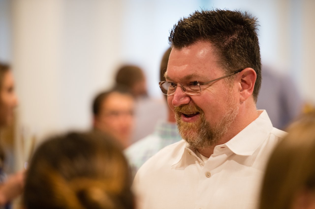 A man in a white shirt smiles while talking with others at an awards dinner.