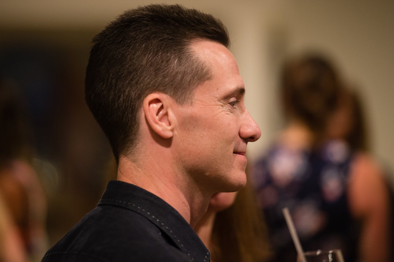 A man in a dark shirt smiles while attending an awards dinner with other guests in the background.