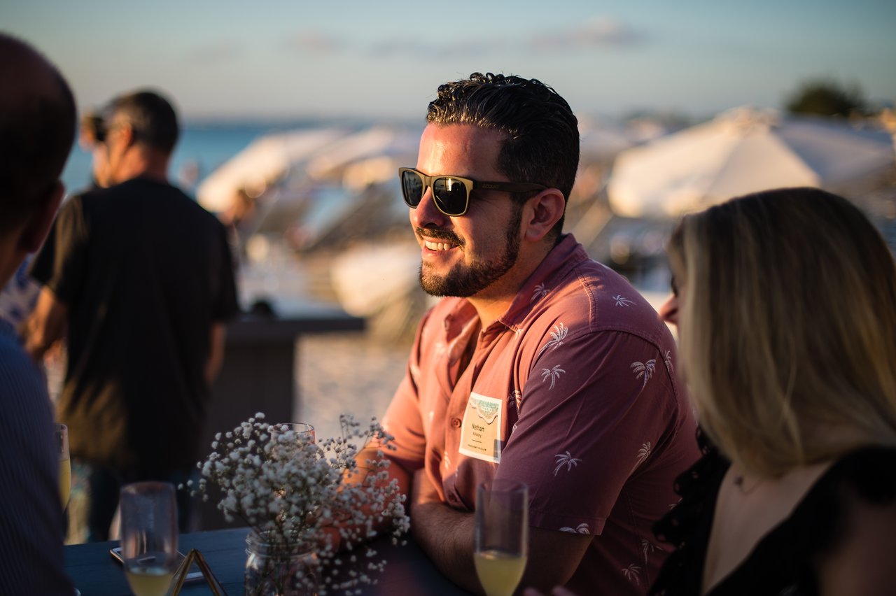 Man in sunglasses and tropical shirt smiles while chatting with others at a beachside company event.