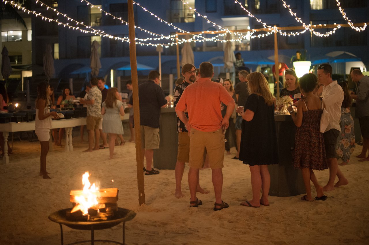 People gather on a sandy beach, chatting and holding drinks under string lights near a fire pit.