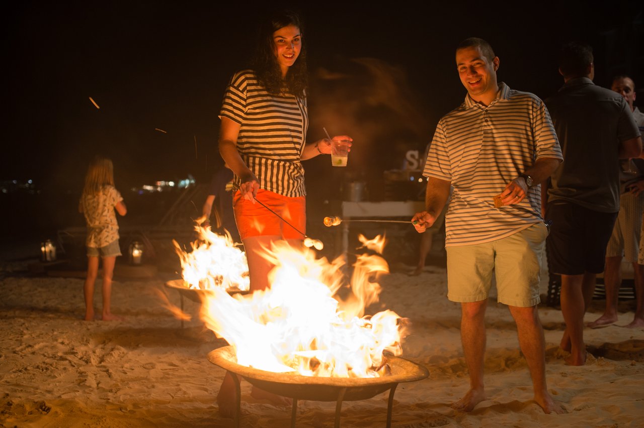 Two people roast marshmallows over a fire pit on a sandy beach at night during a company event.