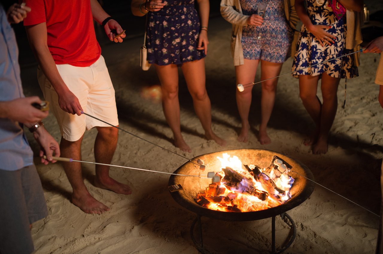 People stand barefoot on the sand, roasting marshmallows over a fire pit during a nighttime beach gathering.