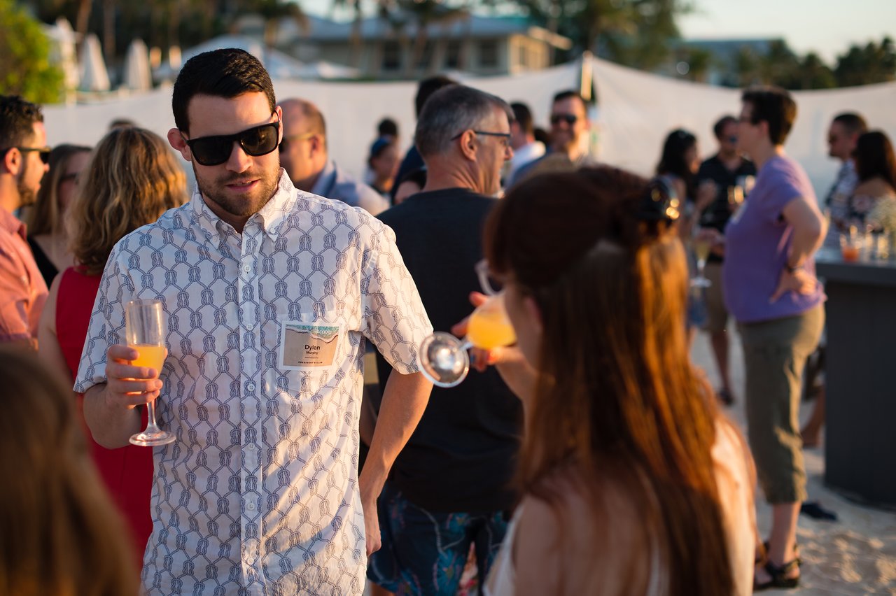 A man in sunglasses holds a drink and talks to a woman at a beach party with colleagues.