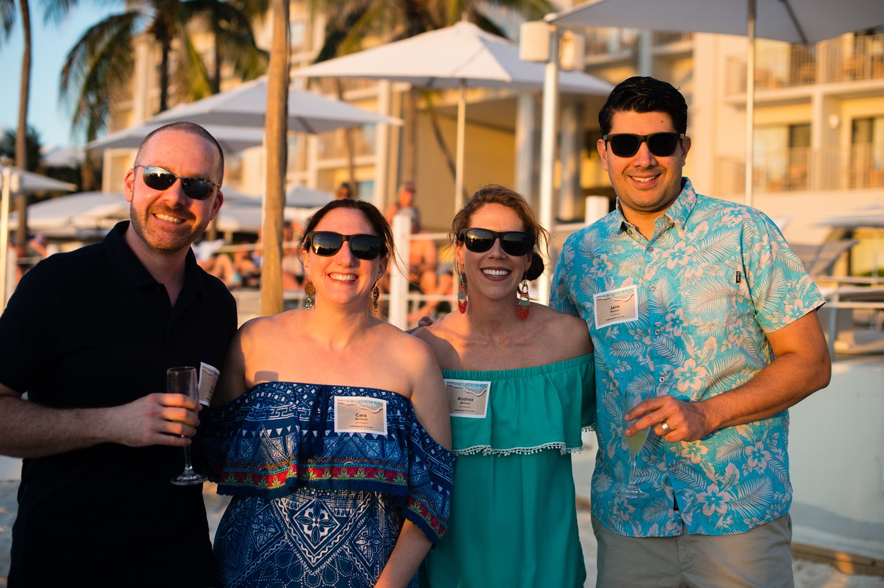 Four people wearing sunglasses smile and hold drinks at a beach party during a company trip in Grand Cayman.