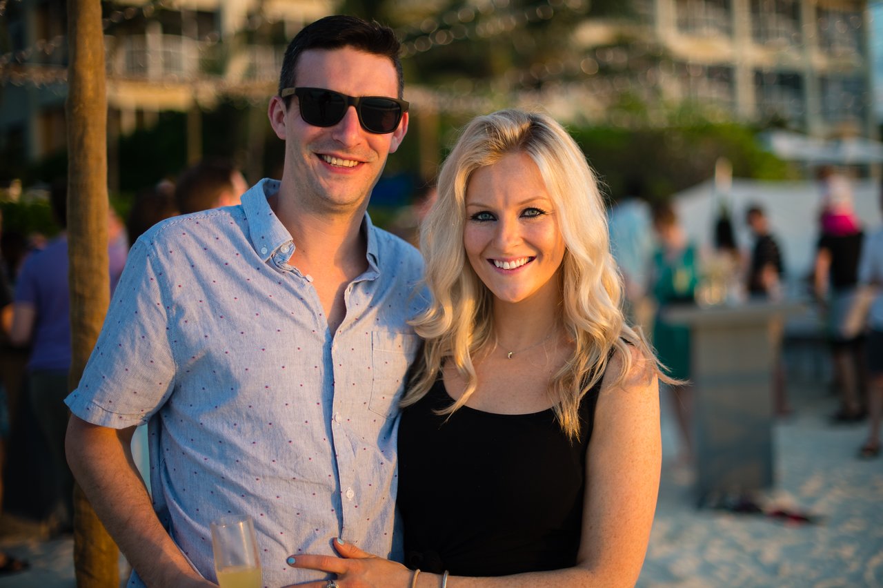 A smiling couple poses at a beach party, with the man holding a drink.