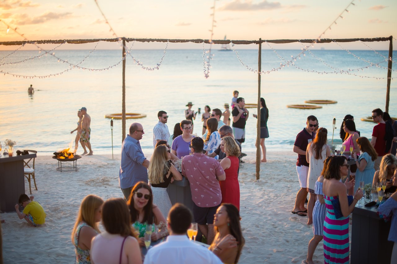 People socializing on a sandy beach, enjoying drinks and conversation during a company trip at sunset.